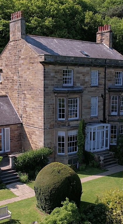 A historic stone building surrounded by manicured gardens and tall hedges, set against a wooded backdrop.