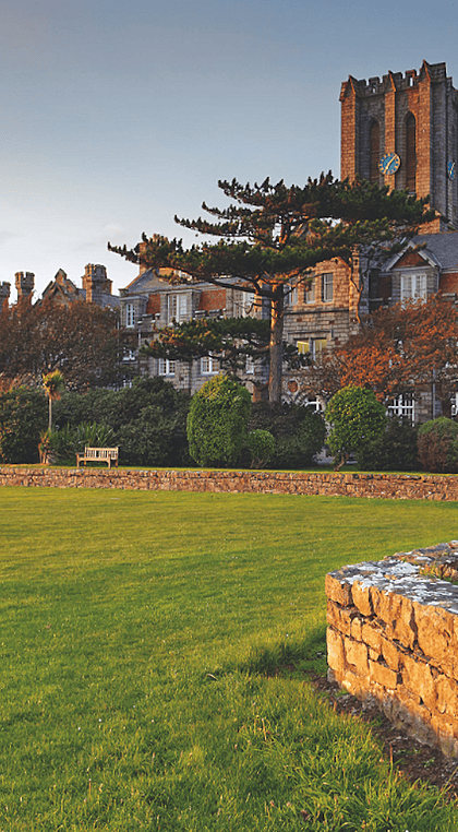 A grand stone school building with a tall tower and surrounding gardens, lit by the warm glow of the setting sun.