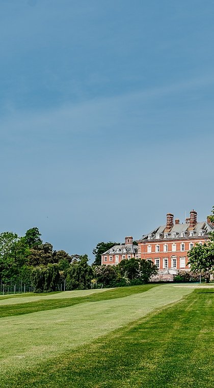 A grand red-brick manor house surrounded by extensive green lawns and tall trees under a clear blue sky.