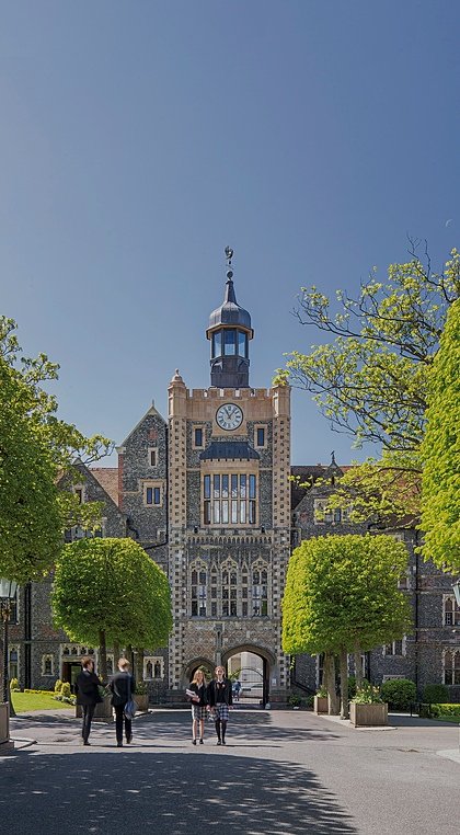 Sunlight school campus with tree-lined path on which pupils amble and a beautiful clocktower in the background