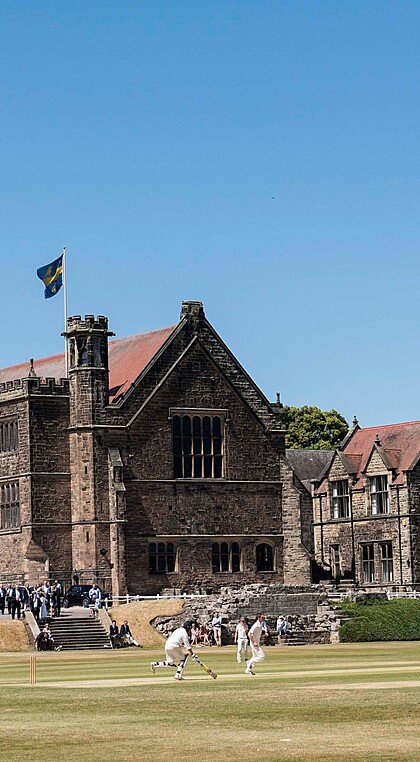 Historic school building with a tall spire and red-tiled roofs, overlooking a cricket match being played on the school grounds.