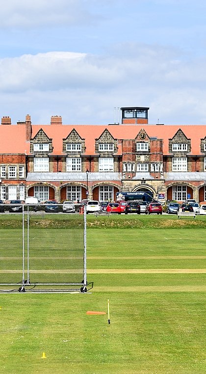 A cricket match is taking place on a large green field in front of a grand red-brick school building with a red-tiled roof and several cars parked outside.