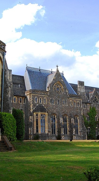 A grand stone school building with Gothic architectural features, including arched windows and detailed stonework, surrounded by green lawns and trees under a partly cloudy sky.