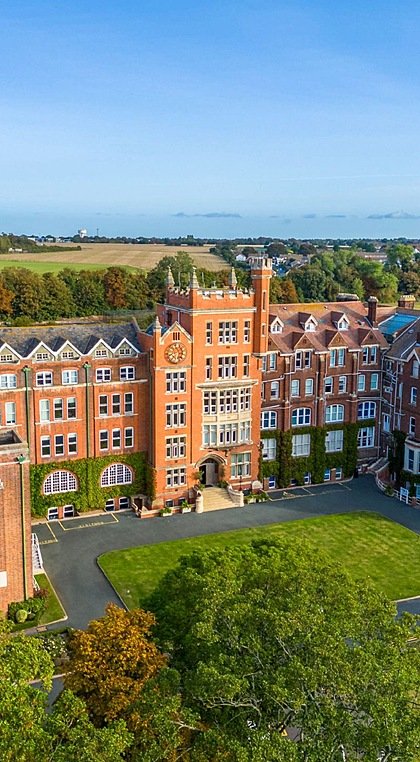 Aerial view of a large red-brick school building with a clock tower, chapel, and green lawns surrounded by trees and open countryside.