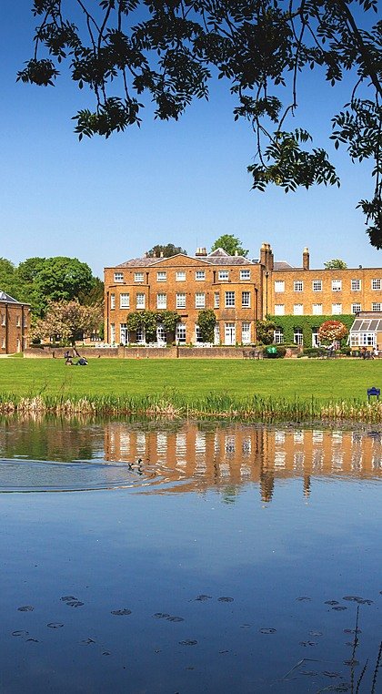 A large yellow-brick manor-style school building with green lawns and a pond in the foreground, surrounded by trees under a clear blue sky