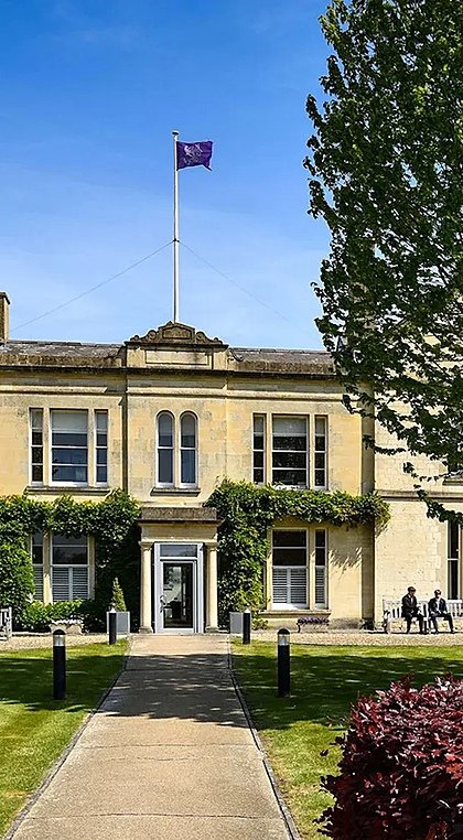 A yellow-brick building with pupils sitting on benches in front of it in the sunshine
