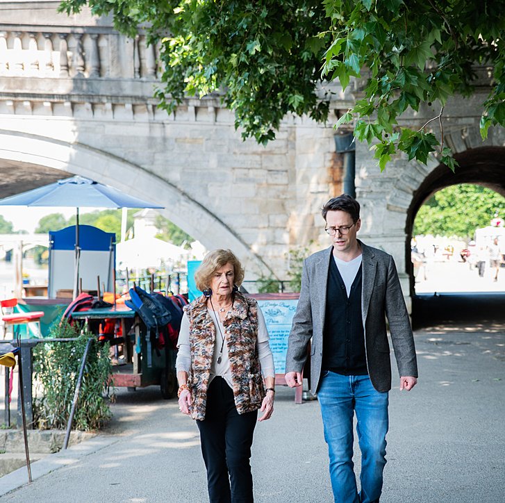 Two people walking together along a riverside path under a stone bridge on a sunny day.