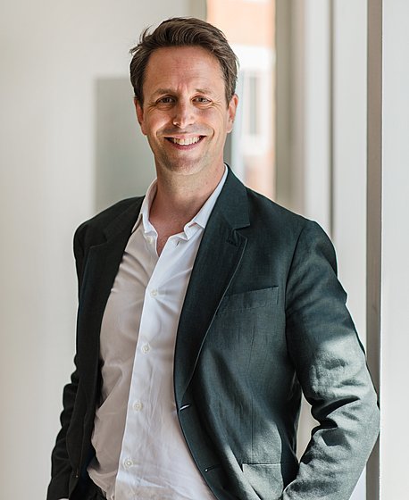 A brunette man in a dark suit with a white shirt is smiling into the camera