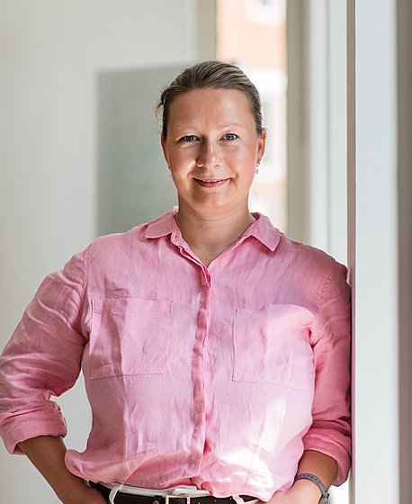 A blond women in a pink shirt and white jeans is leaning against a window and smiling into the camera