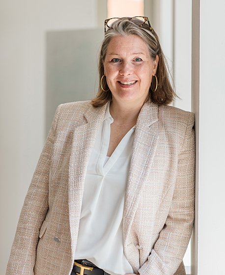 A blond women with her glasses on her head in a white blouse and beige blazer is leaning against a window and smiling into the camera