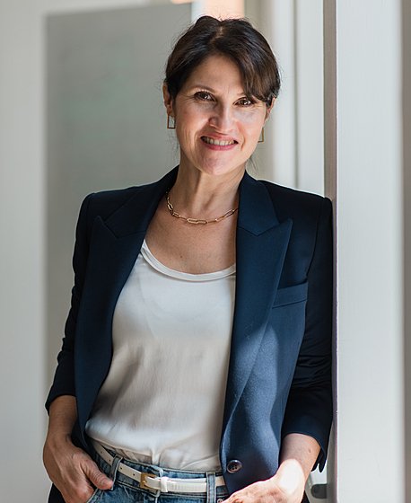 A brunette women in a navy blazer white T-Shirt and jeans is leaning against a window and smiling into the camera