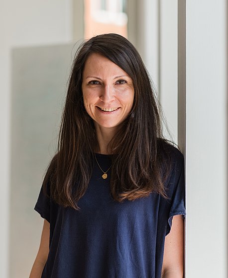 A woman with long brown hair wearing a navy blue shirt smiles while standing against a white wall in a brightly lit room.