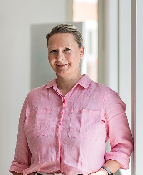 A blond woman in a pink shirt and white jeans is smiling into the camera