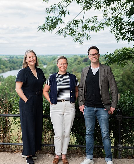 two women and a man are standing together under a tree in front of a nice vista, smiling into the camera