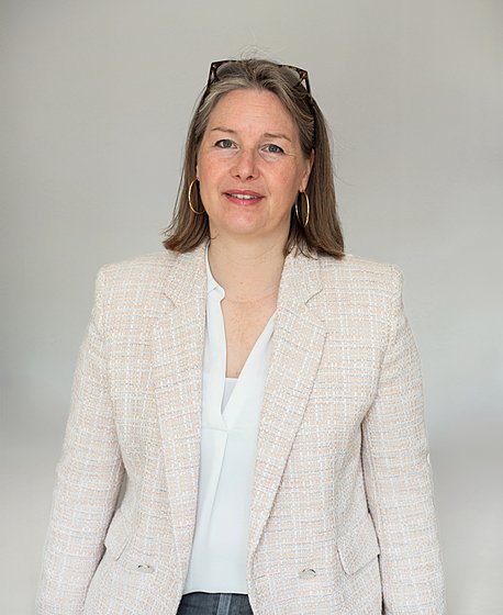 A blond women wearing her glasses on the head in a beige blazer and a white blouse is looking into the camera