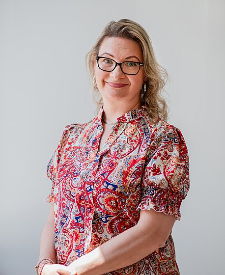 A blond women with glasses on in a patterned blouse is smiling into the camera