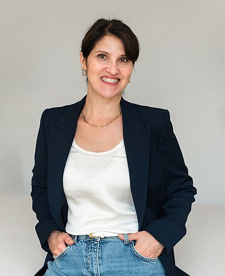 A brunette women in a navy blazer, a white t-shirt and a jeans is smiling into the camera