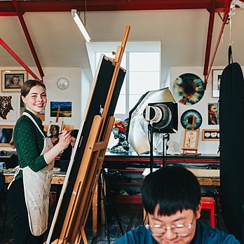 Two children in an art atelier painting, with lots of paintings on the wall