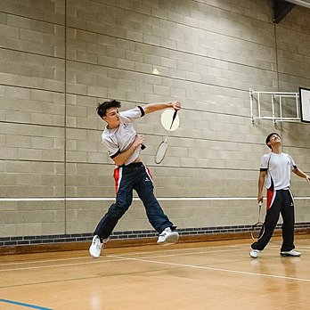 Two students playing badminton indoors, one jumping mid-air to hit the shuttlecock.