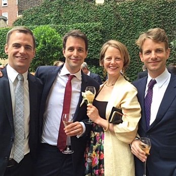 Four people dressed in formal wear smiling at an outdoor event, holding glasses of champagne.