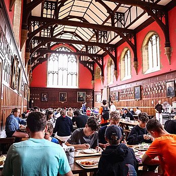 Pupils eating and chatting in a grand dining hall with wooden panelled walls and high arched windows.