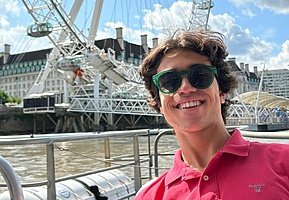 A person wearing sunglasses smiles by the river with a large Ferris wheel and city buildings in the background.