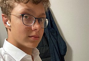 A boy wearing glasses, a white shirt, and wireless earbuds looks into the camera indoors.