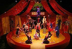 A group of performers in a circus-themed stage production with vibrant red curtains and spotlights.