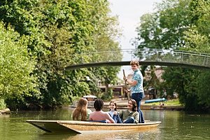 Students enjoying a boat ride on a calm river surrounded by green trees and a bridge overhead.