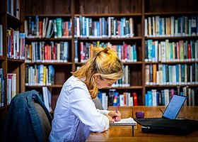 A student learning making notes on a notepad in front of an open laptop in a library