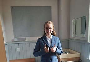 a blonde girl smiling in her school uniform gives two thumbs up into the camera