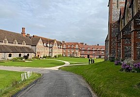 Expansive school grounds with red-brick and flint buildings surrounding a neatly mowed lawn under a cloudy sky.