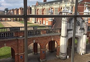 View through a large window of a grand courtyard surrounded by ornate red-brick and stone buildings.