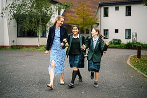 An adult woman walks with two pupils in school uniform along a paved path on campus.