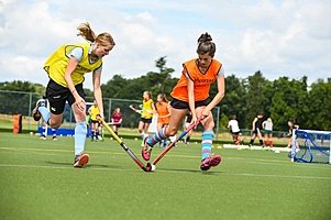 Two students play field hockey on a green astroturf pitch, competing for the ball during a sunny practice session.