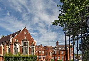 Traditional red-brick school buildings framed by wrought-iron gates and leafy trees under a blue sky.