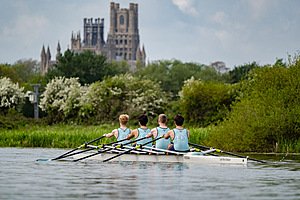 Four boys in a rowing boat on the water with a beautiful old School building in the background