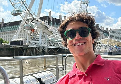 A person wearing sunglasses smiles by the river with a large Ferris wheel and city buildings in the background.