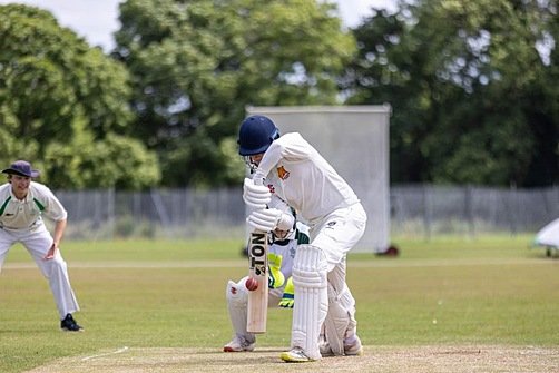 A cricket player in white uniform batting during a match, with a teammate and wicketkeeper visible in the background.