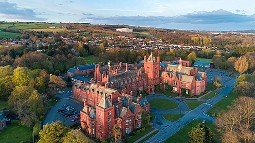 Aerial view of a large red-brick school complex set among trees with a town and rolling hills in the background.