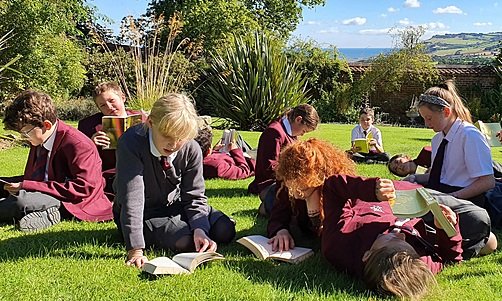 Students in school uniforms reading books while sitting and lying on the grass in the sunshine.