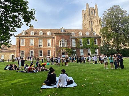 Students gather and relax on a lawn in front of a historic red-brick building covered partly with ivy, with a tall cathedral-like tower in the background.