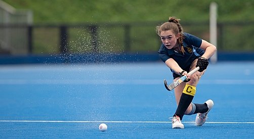 A girl hits the Hockey ball with her stick outside on a blue pitch