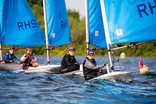 Pupils sailing small boats with bright blue sails on a calm lake, concentrating as they steer and adjust their ropes.