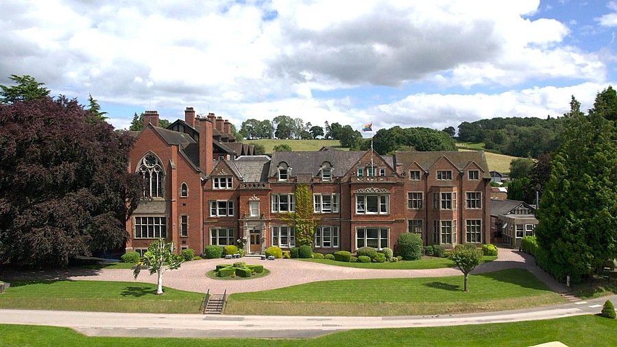 A large red-brick school building surrounded by lawns and trees, with a grand entrance and countryside hills in the background.