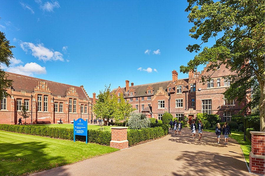 Wide-angle shot of a school campus with green lawn and pupils on foreground and red-brick school buildings in back