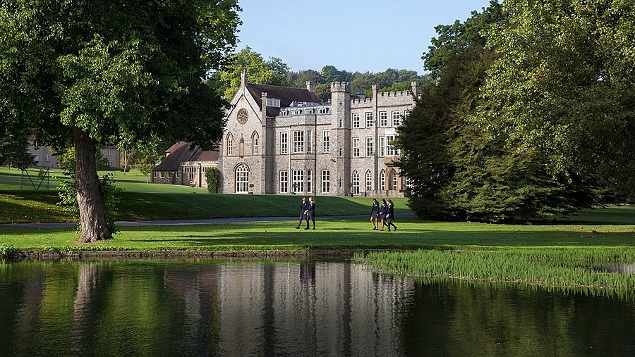 An imposing, grey manor house building with a lake and trees in the foreground with girls going for a walk