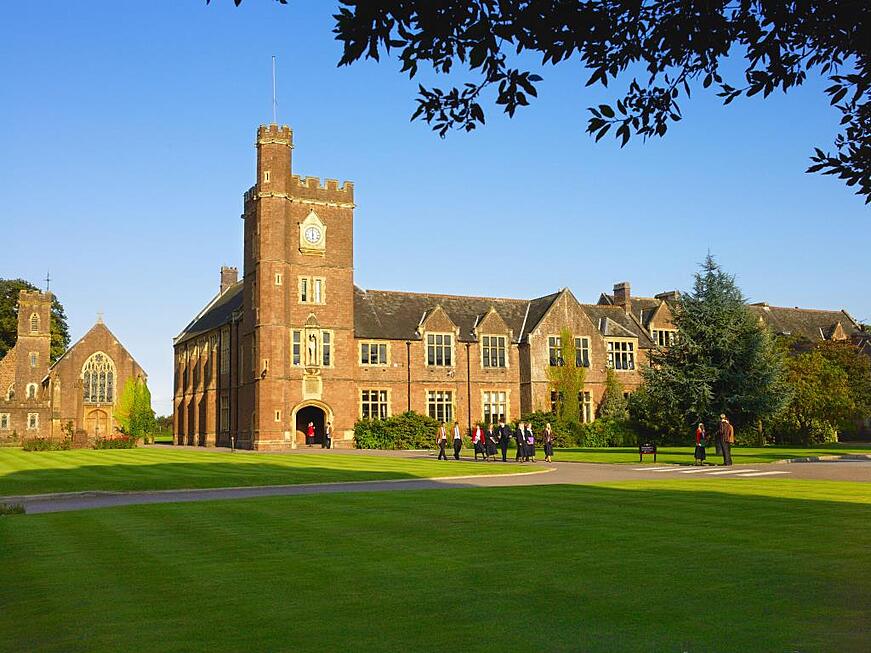 Red-brick school building with clock tower and students walking across a green lawn.