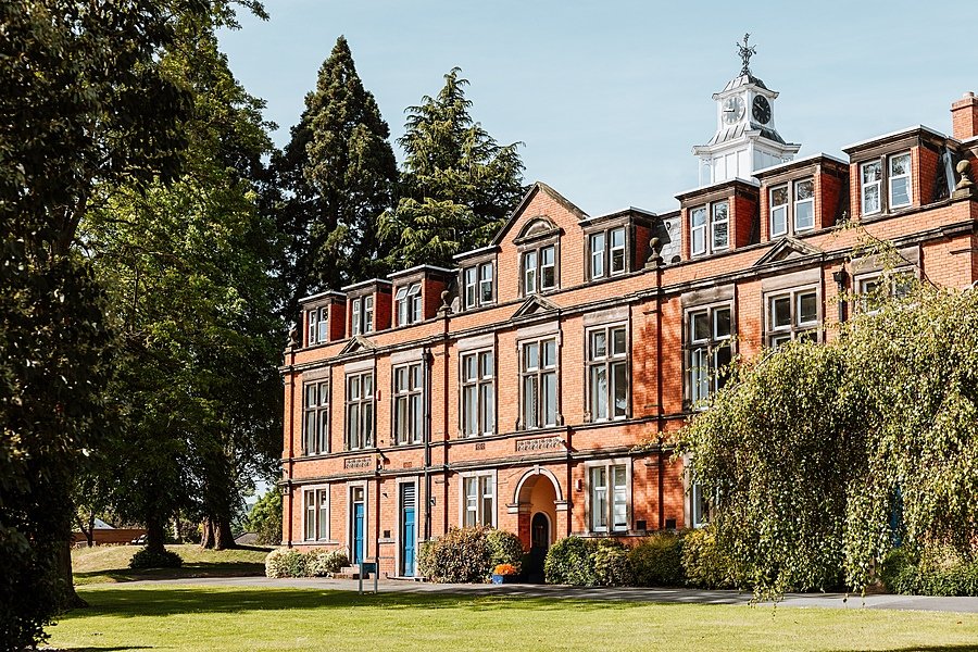 Historic red-brick school building with large windows and a clock tower surrounded by trees.