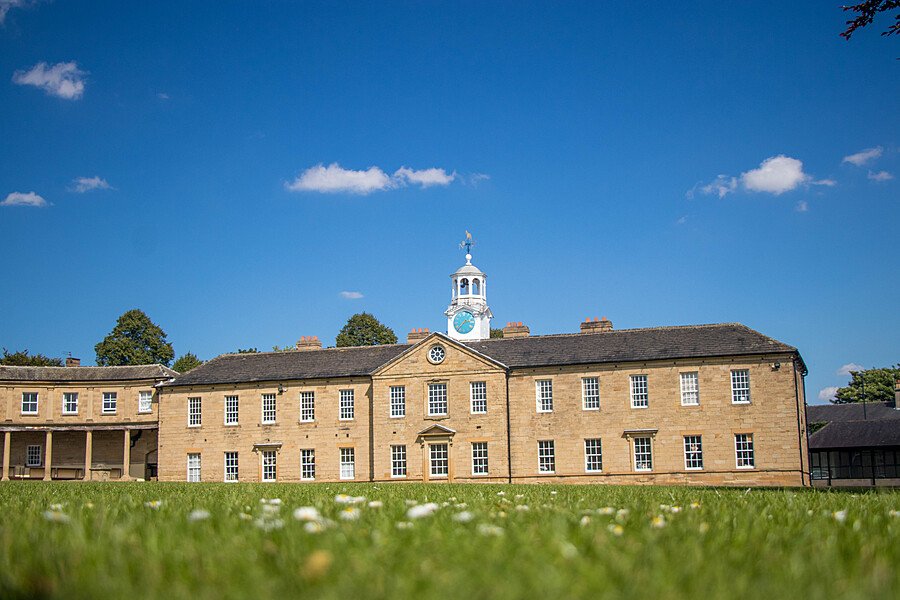 A historic sandstone school building with a clock tower and cupola, set against a bright blue sky and surrounded by a well-kept lawn.